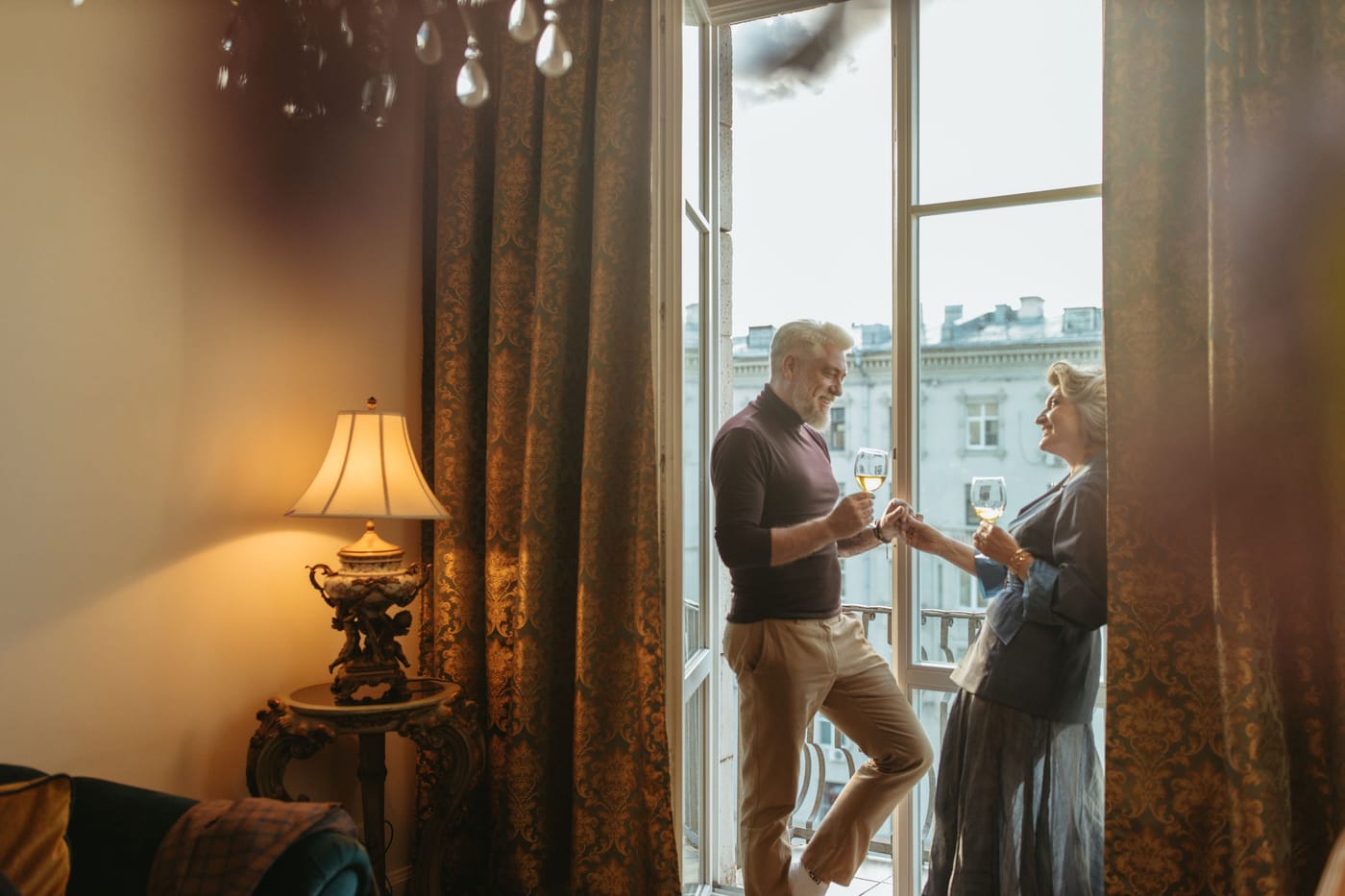 Ein Paar mit silbergrauem Haar stößt am hohen Fenster eines eleganten Altbauzimmers mit Sekt an.